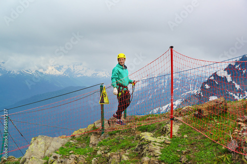 A young man in a helmet and with rescue ropes should on top of the mountain at the cliff