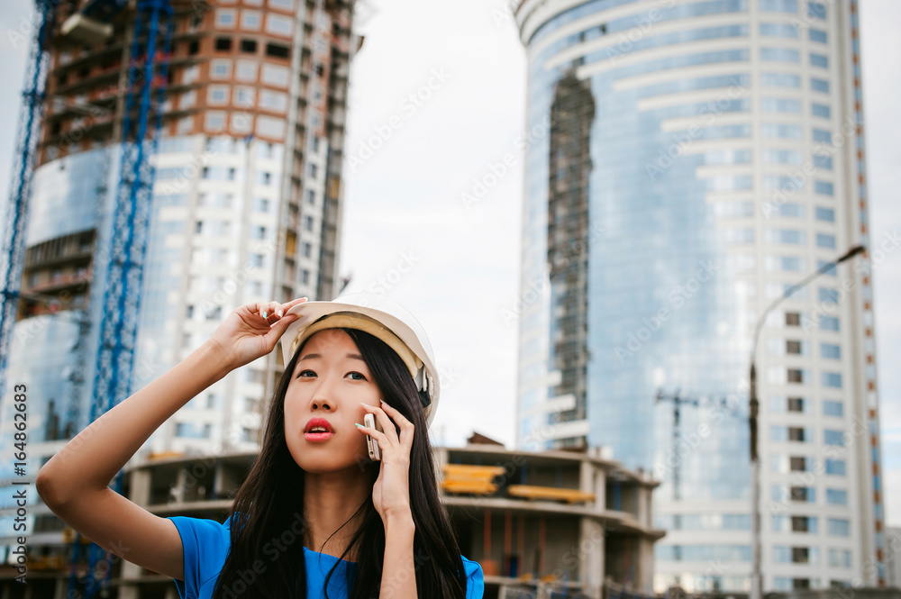 Fototapeta premium Asian woman in a blue dress and white protective helmet. engineer supervises inspection and control of works at construction site. Business telephone conversations. Communication via smartphone