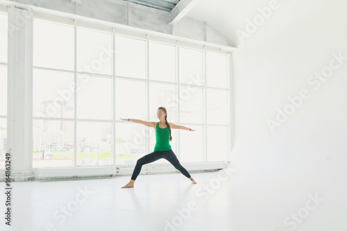 Beautiful smiling happy young woman working out indoors, doing yoga exercise near floor window with city view