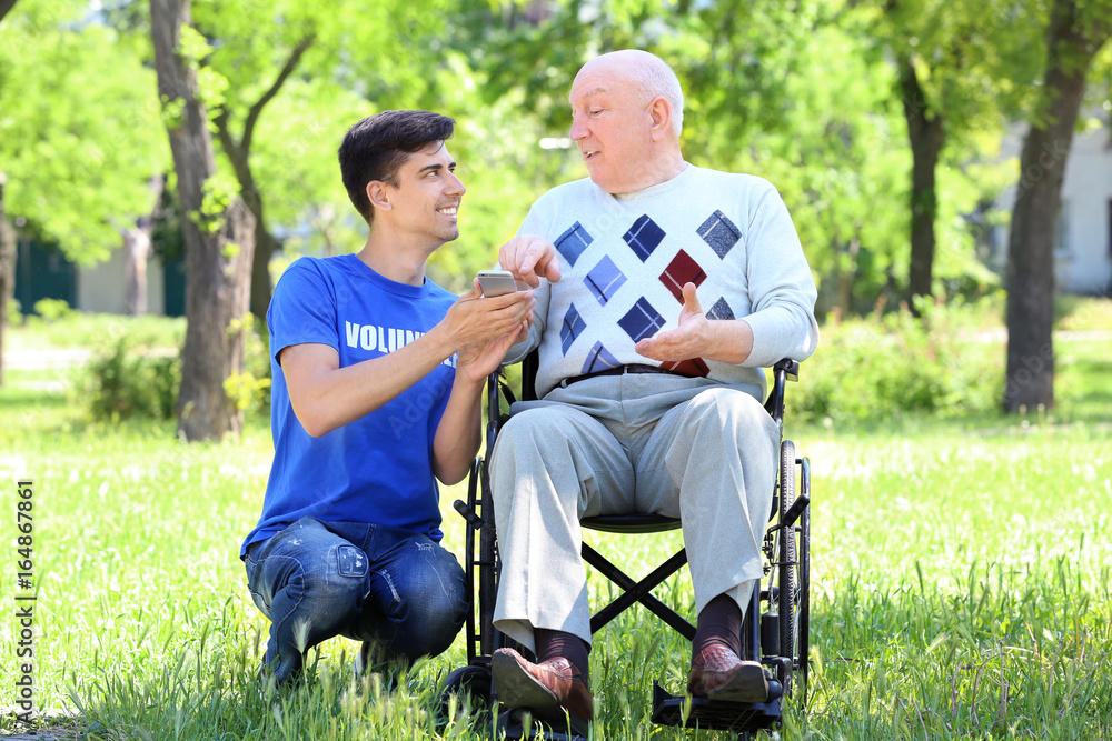 Young male volunteer and senior man on wheelchair with mobile phone outdoors Young male volunteer and senior man on wheelchair with mobile phone outdoors