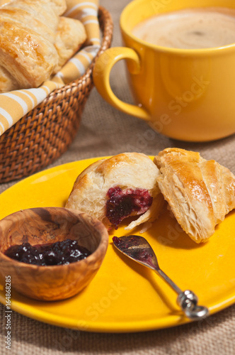 A fresh, baked croissont ripped open and a raspberry jam spread  with an artisan knife on a round, yellow plate with a cup of cappuccino and a basket of buns photo