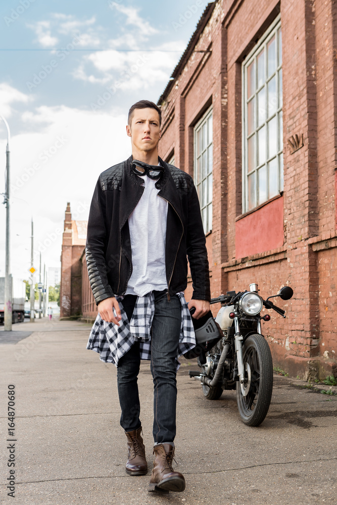 Handsome rider biker man in black leather jacket, jeans and helmet walk  away from classic style cafe racer motorcycle. Bike custom made in vintage  garage. Brutal fun urban lifestyle. Outdoor portrait. Stock