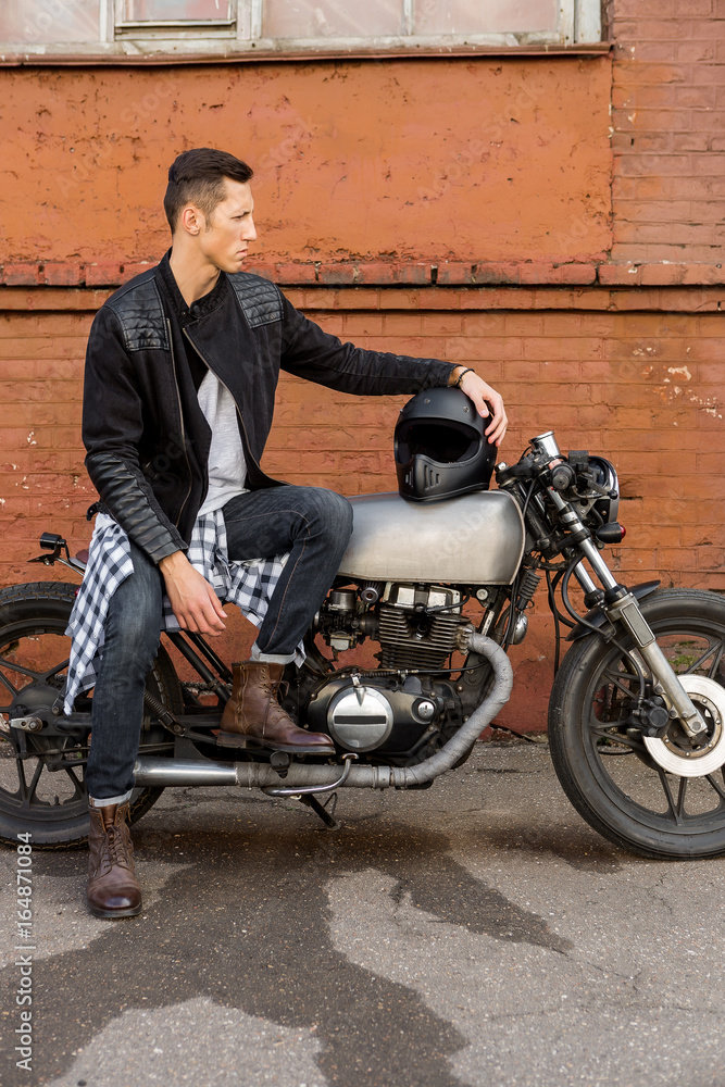Handsome rider biker guy in leather jacket sit on classic style cafe racer motorcycle and hold hand on black helmet. Bike custom made in vintage garage. Brutal fun urban lifestyle. Outdoor portrait.
