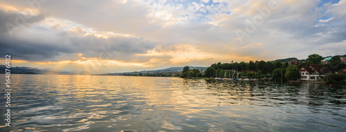 Panoramic summer view of boat cruise excursion landscape on Zurichsee with beautiful sunset shining light through clouds reflected the golden light on the water surface as a background., Rapperswil.