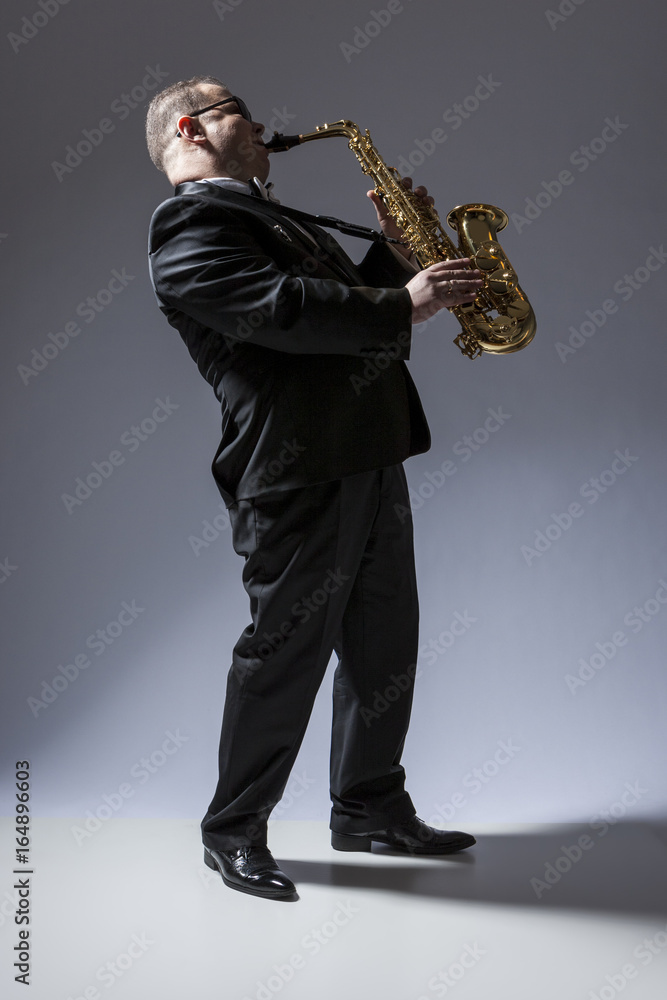 Naklejka premium Music Concepts. Full Length Portrait of Mature Expressive Caucasian Saxophone Player in Sunglasses Playing the Saxophone in Studio Environment.