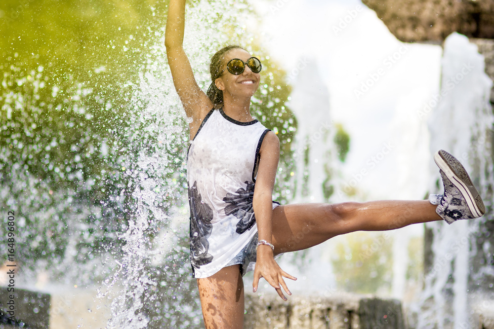 Fototapeta premium Teenager Concepts. Portrait of Smiling Funny African American Teenager Girl with Dreadlocks Enjoying in Fountain Outdoors.With Hand and Leg Lifted