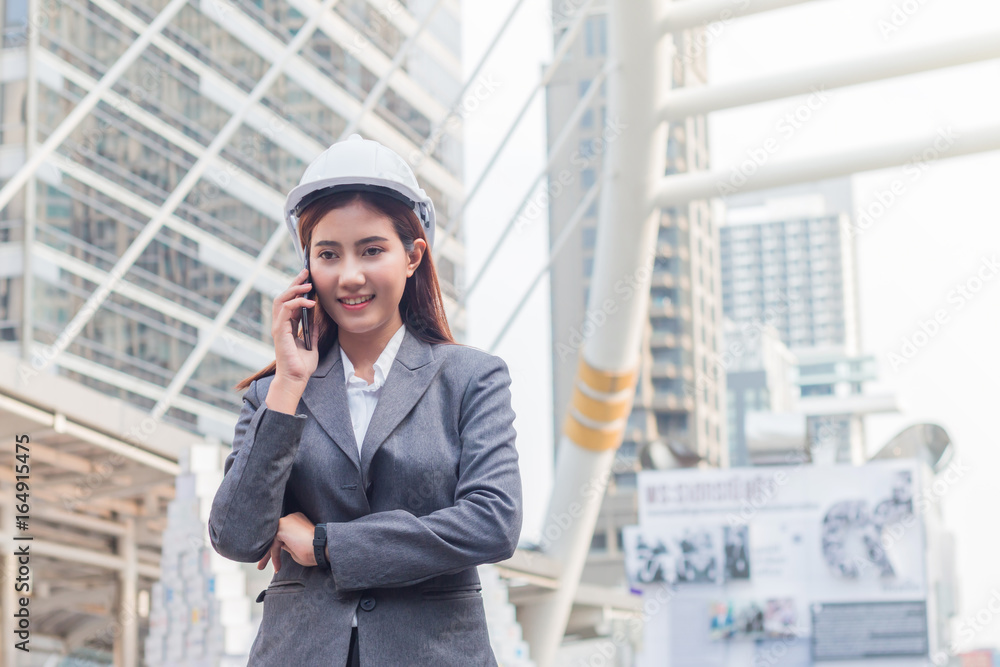 Portrait of beautiful young engineer woman wear a white safety helmet. She are smile and talking on smartphone with commitment to success with building background