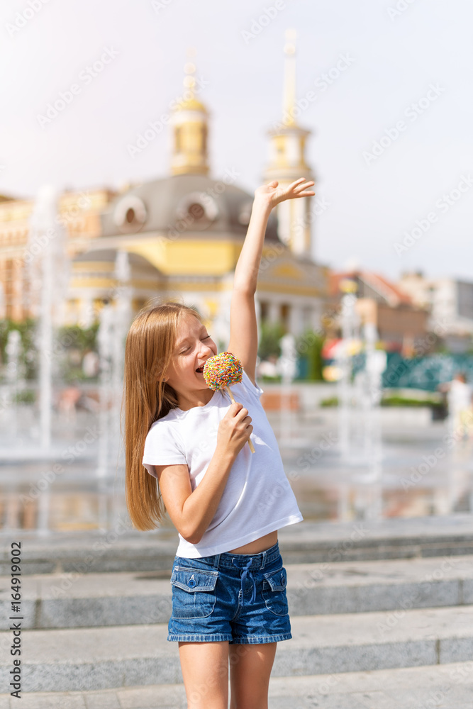 Cute little girl eating candy apple, singing pretending the candy is microphone at fair in amusement park.