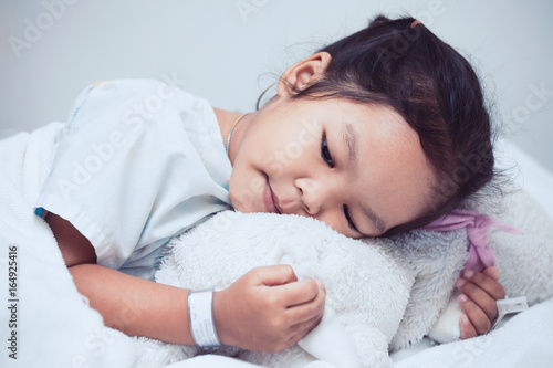 Sick asian child girl is lying in the bed and hugging her doll in the hospital