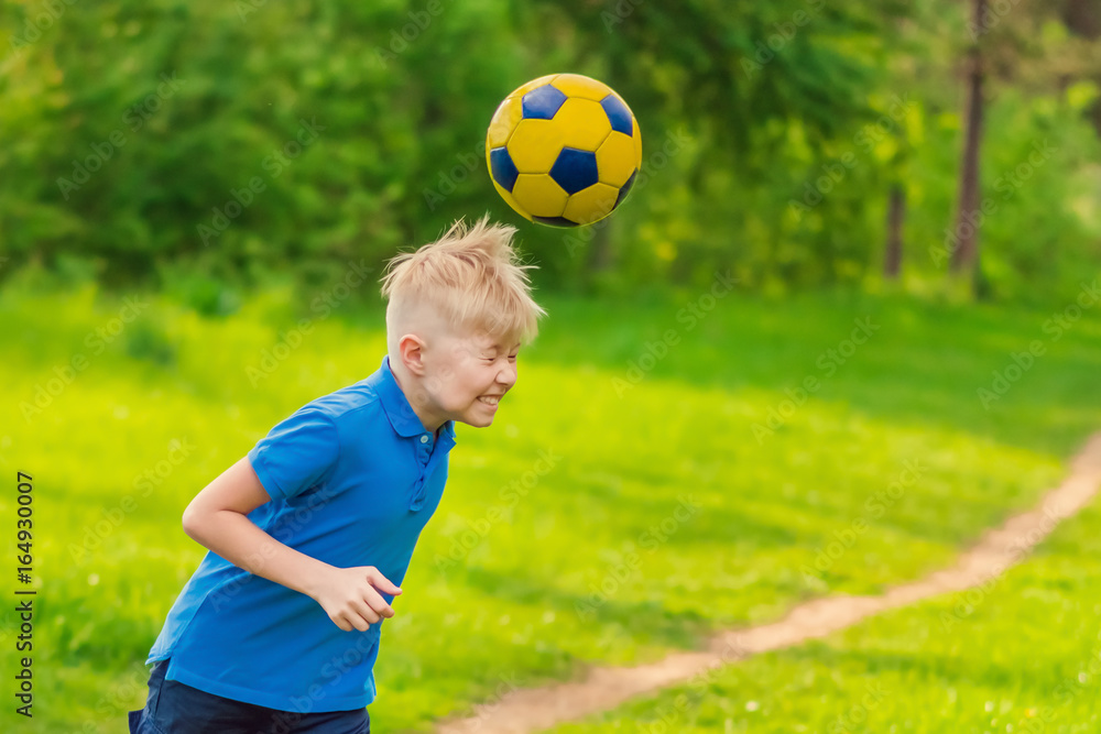 Blond boy in a white T-shirt boy beats his head a soccer ball in the park Blond boy in a white T-shirt boy beats his head a soccer ball in the park
