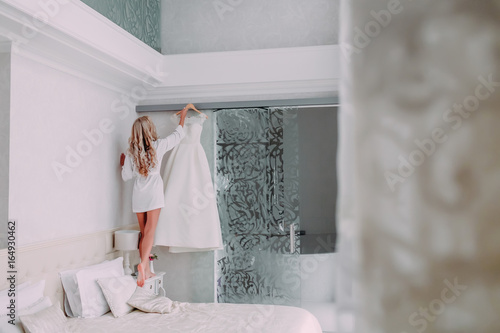 Bride in silk robe takes her dress from the cornice. Morning preparation before wedding