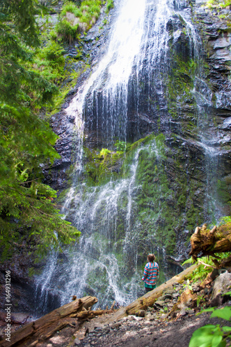 One young girl standing with her back turned, looking at remote desolate Yalynskyi waterfall with water drops falling against rocky slope. Verdant plants, trees, logs. Carpathian mountains, Ukraine photo