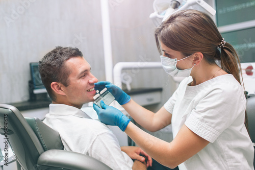 Portrait of a female dentist and young happy male patient in a dentist office. Man checking and selecting colour of the teeth