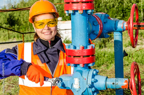 Woman worker in the oilfield near wellhead, wearing orange helmet and work clothes. Industrial site background. Oil and gas concept.