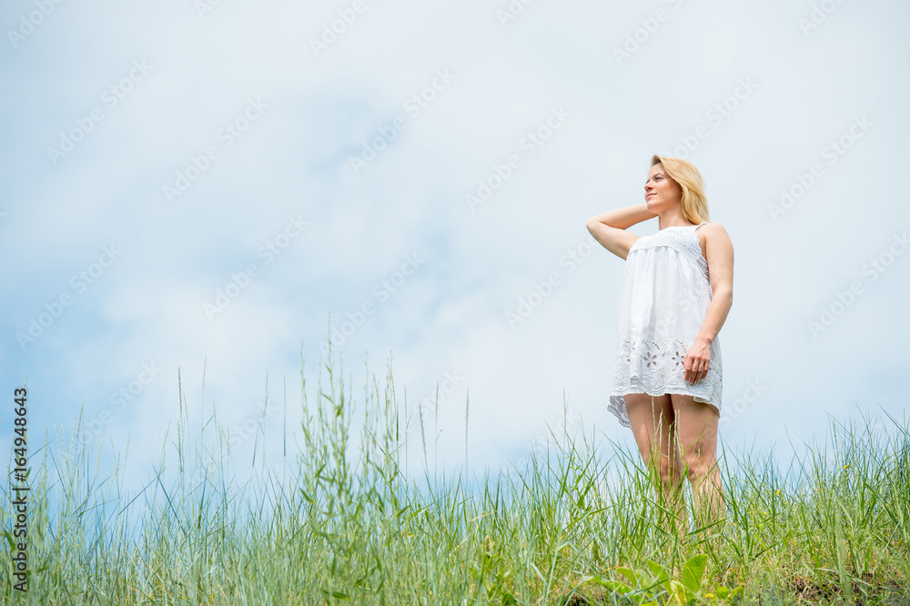 Beautiful girl in short white dress, posing on a meadow on warm sunny day. Sky and grass background