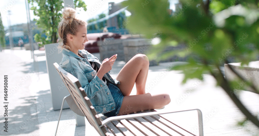 Beautiful young woman wearing denim jacket typing on phone in a city park during sunny day. Pretty young girl sitting on wooden bench with phone. 