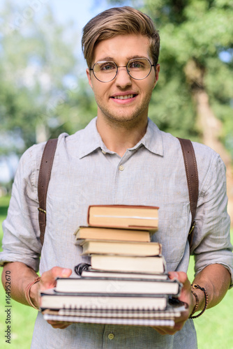 habdsome young man in eyeglasss holding pile of books and smiling at camera photo