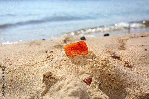 Fototapeta Naklejka Na Ścianę i Meble -  Amber. A red piece of Baltic amber on a sandy beach against a background of sea waves. Natural mineral amber in the sand lit by the sun. Frozen resin as an ornament in jewelry production.
