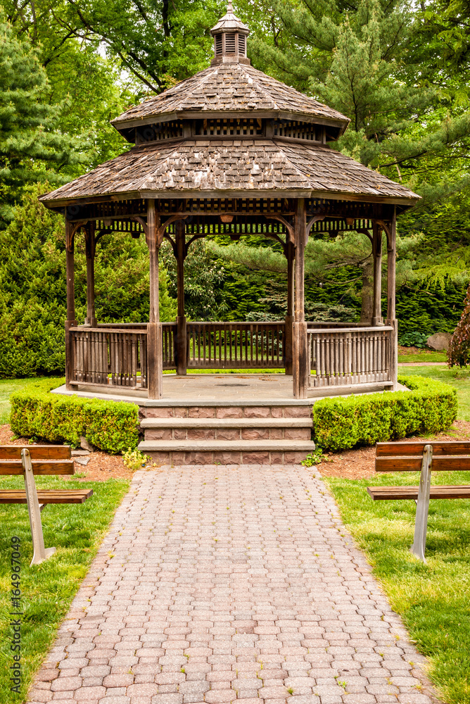 Front of gazebo with wood and steel benches in front. Outdoor gazebo with shrubs and grass. Shady retreat in backyard. Outdoor rotunda summerhouse. Outside platform.