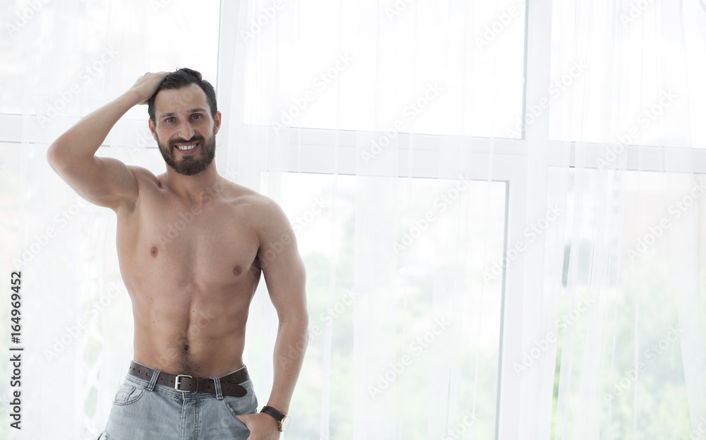 Sexy fashion portrait of a hot bearded male model in stylish jeans with muscular body posing in modern interior setting with window light Sexy fashion portrait of a hot bearded male model in stylish jeans with muscular body posing in modern interior setting with window light