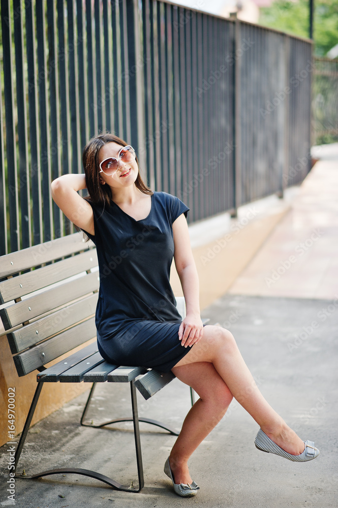 Brunette girl at black dress on sunglasses sitting on bench and posing at street of city.