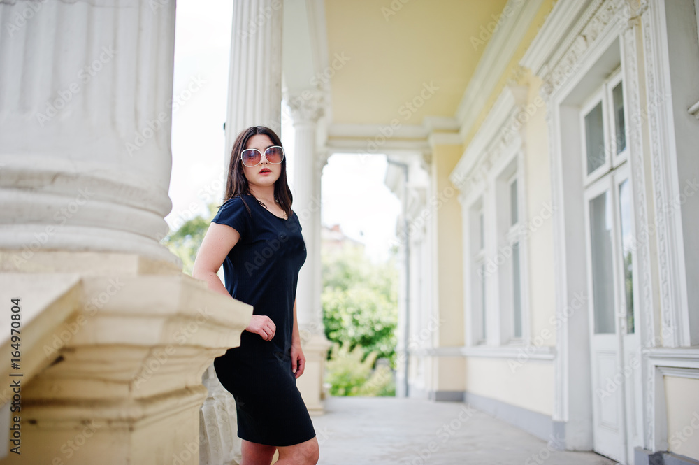 Brunette girl at black dress, sunglasses posed against old vintage house, at street of city.