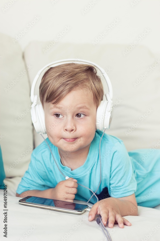 3 year old boy sits on the couch and listens to music. Little cute boy watching cartoons on the phone. Little boy. 3 year old boy sits on the couch and listens to music. Little cute boy watching cartoons on the phone. Little boy.