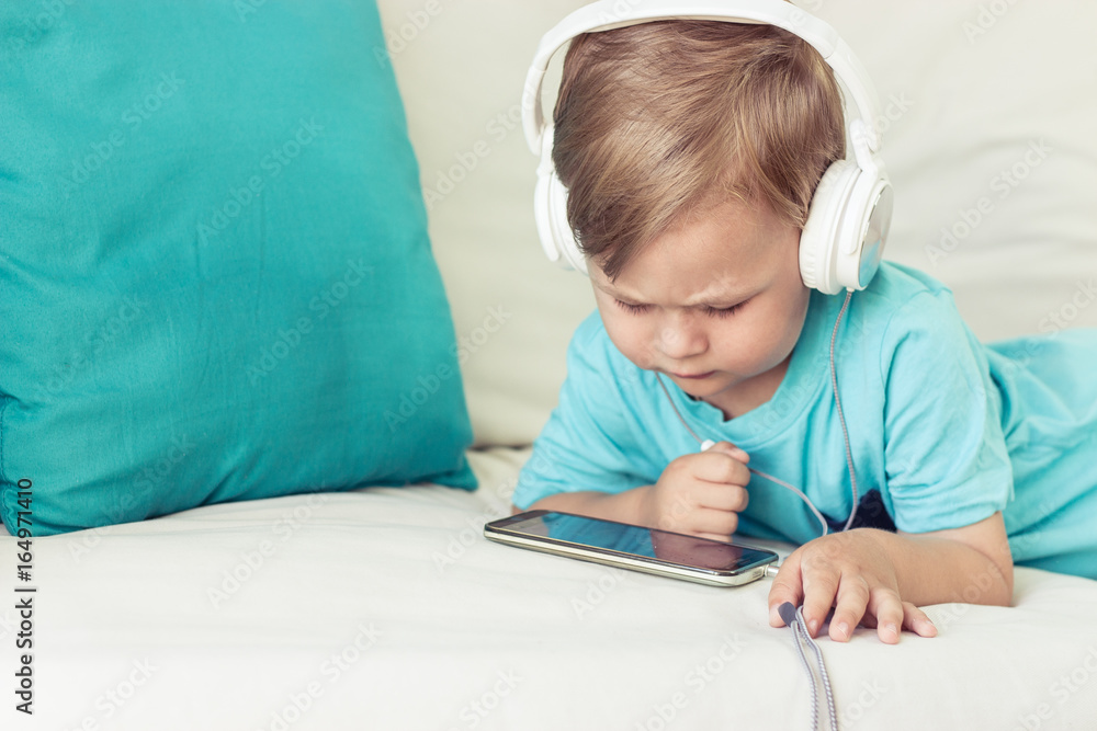 3 year old boy sits on the couch and listens to music. Little cute boy watching cartoons on the phone. Little boy. 3 year old boy sits on the couch and listens to music. Little cute boy watching cartoons on the phone. Little boy.