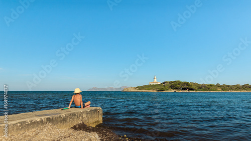 Child seated on a jetty looking at the sea and a lighthouse on a islet, Alcudia in Balearic Islands photo