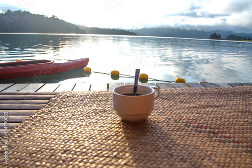 White cup of hot coffee on wood net and morning lighting behind to see lake view for refresh time
