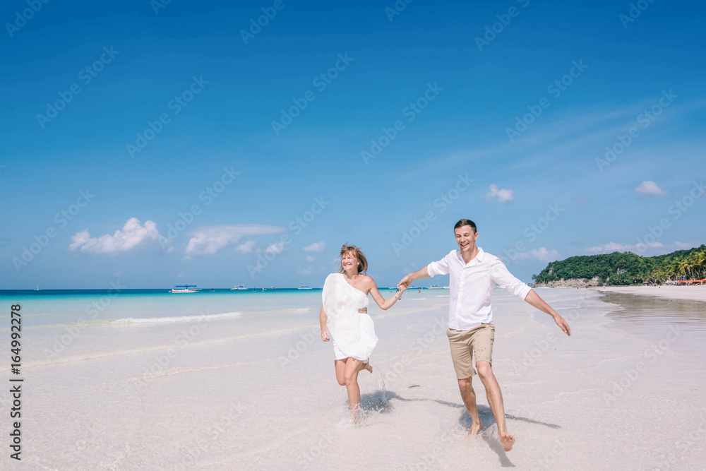 Active couple having fun and running on a white sandy beach with blue sky and turquois water on the background. Men and women are happy and laughing together