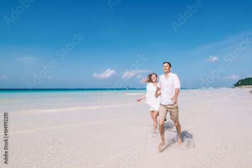 Active couple having fun and running on a white sandy beach with blue sky and turquois water on the background. Men and women are happy and laughing together