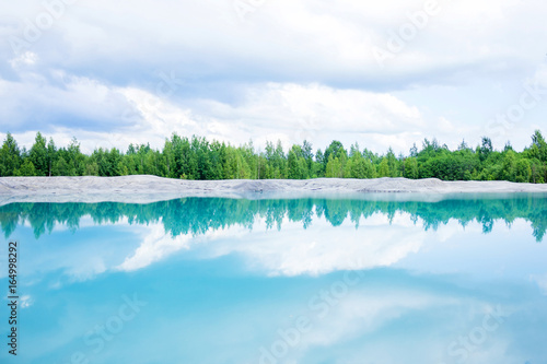 Fototapeta Naklejka Na Ścianę i Meble -  A lake with saturated blue water next to an abandoned mine. Young green trees in the background.  Pollution of environment as an ecological problem