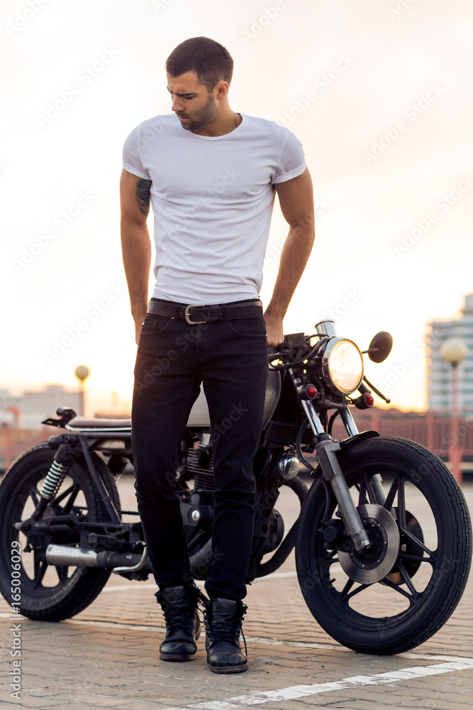 Obraz premium Mężczyzna przystojny jeździec sportowy rowerzysta w biały t-shirt puste odejść od klasycznego motocykla racer kawiarnia o zachodzie słońca. Vintage rower wykonany w garażu. Brutalny miejski styl życia. Portret na zewnątrz.
