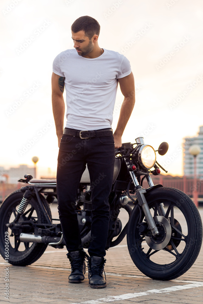 Sporty biker handsome rider man in white blank t-shirt walk away from classic style cafe racer motorbike at sunset. Vintage bike custom made in garage. Brutal urban lifestyle. Outdoor portrait. Sporty biker handsome rider man in white blank t-shirt walk away from classic style cafe racer motorbike at sunset. Vintage bike custom made in garage. Brutal urban lifestyle. Outdoor portrait.