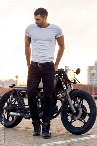 Sporty biker handsome rider man in white blank t-shirt walk away from classic style cafe racer motorbike at sunset. Vintage bike custom made in garage. Brutal urban lifestyle. Outdoor portrait.