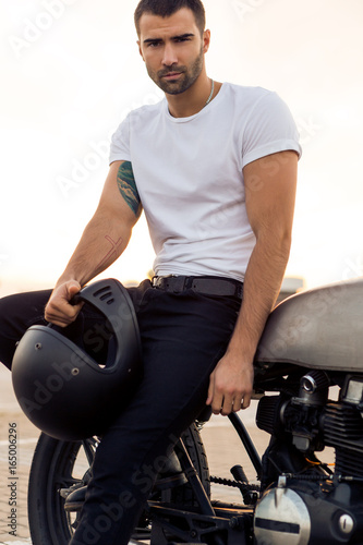 Close up of a handsome rider male in white blank t-shirt near classic style cafe racer motorcycle at sunset. Bike custom made in vintage garage. Brutal urban lifestyle. Outdoor portrait.