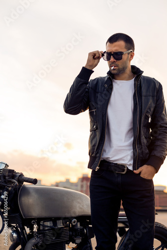 Closeup of a handsome rider guy with beard and mustache in black biker jacket take off fashion sunglasses smoking cigaret near classic style cafe racer motorbike at sunset. Brutal fun urban lifestyle.