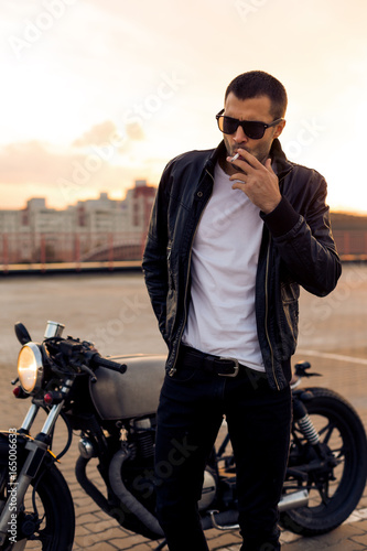 Beautiful happy rider man with beard and mustache in black biker jacket, jeans and fashion sunglasses smoking cigaret near classic style cafe racer motorbike at sunset. Brutal fun urban lifestyle.