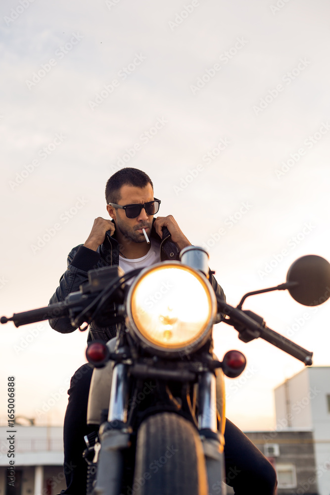 Handsome rider man with beard and mustache smoking cigaret and correct biker jacket sit on classic style cafe racer motorbike at sunset. Bike custom made in vintage garage. Brutal fun urban lifestyle. Handsome rider man with beard and mustache smoking cigaret and correct biker jacket sit on classic style cafe racer motorbike at sunset. Bike custom made in vintage garage. Brutal fun urban lifestyle.