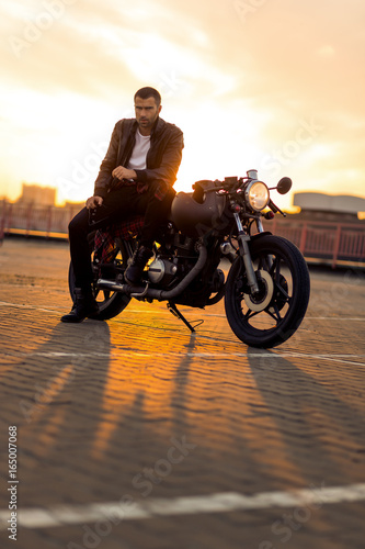 Handsome rider man with beard and mustache in black leather biker jacket sit on classic style cafe racer motorcycle rooftop at sunset. Bike custom made in vintage garage. Brutal fun urban lifestyle.