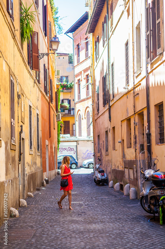Fototapeta Naklejka Na Ścianę i Meble -  Girl in red dress walking on Rome street. Trastevere, Rome, Italy. Young girl. Old street in Rome. Pretty girl around the corner. Rome architecture. Blonde hair girl