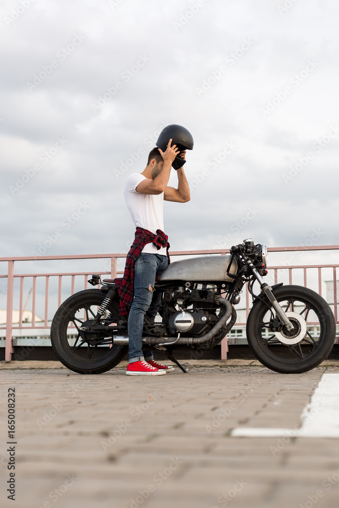 Handsome rider man with beard and mustache posing while put on black moto  helmet near classic style biker cafe racer motorcycle. Bike custom made in  vintage garage. Brutal fun urban lifestyle. Stock