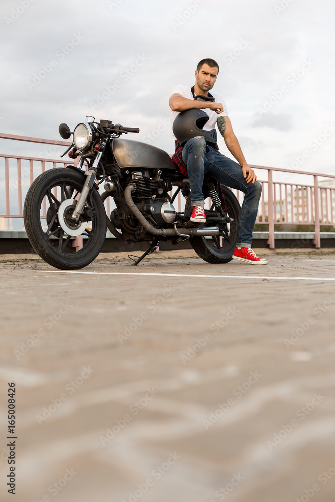 Foto Stock Rider guy with beard and mustache in red sneakers sit on classic  style biker cafe racer motorcycle with black helmet and look to camera.  Bike custom made in vintage garage.