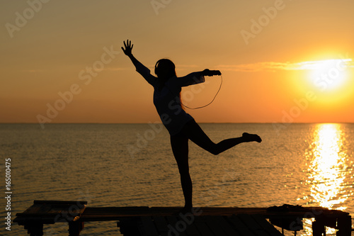 A girl in lingerie and shirt listening music on headphones on a pier by the sea at sunset