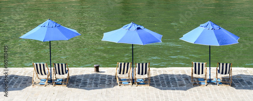 Three blue parasols with blue and white striped deck chairs in the sun on the wharf of the river Seine with green water.