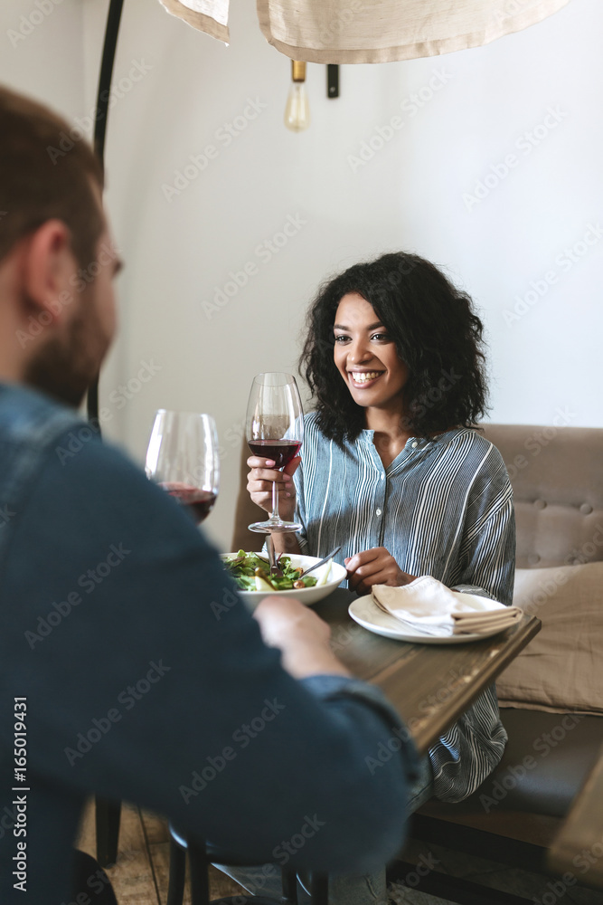 Young couple sitting in restaurant and drinking red wine. Pretty African American girl eating salad and drinking wine at cafe. Smiling lady with dark curly hair having dinner with friend in restaurant Young couple sitting in restaurant and drinking red wine. Pretty African American girl eating salad and drinking wine at cafe. Smiling lady with dark curly hair having dinner with friend in restaurant
