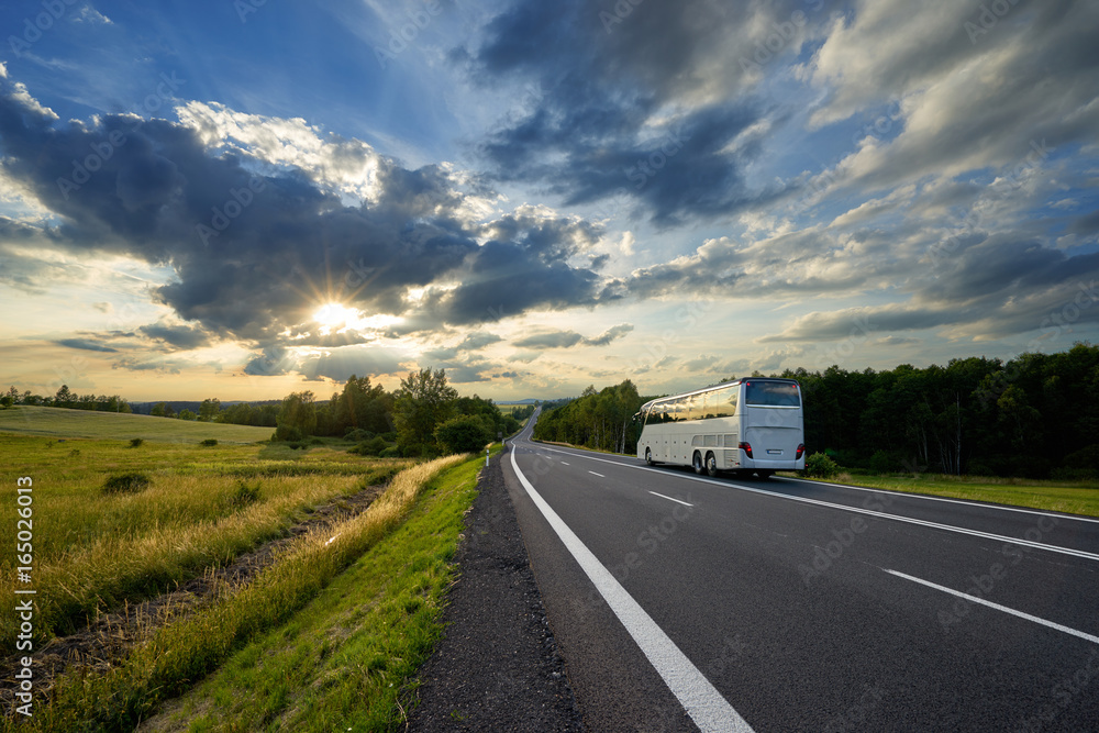 Bus traveling on the asphalt road in a rural landscape at sunset with dramatic clouds