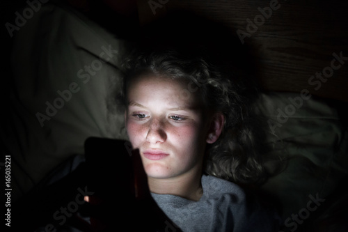 Close up of a teenage girls face lying on a pillow in bed lit up by the light from a cellphone