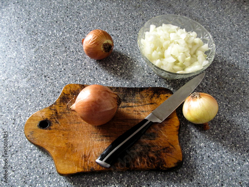 A bowl with chopped onions, three whole onions and a knife on a cutting board - the cooking stage. Gray spotted